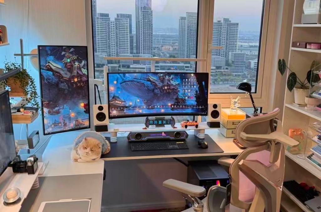 Multi-screen home workspace with computer monitors, a camera, speakers and a cat beside a large window overlooking high-rise buildings.